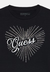 Black organic cotton t-shirt featuring a central "Guess" logo in silver with radiating white rhinestones in a heart shape pattern.