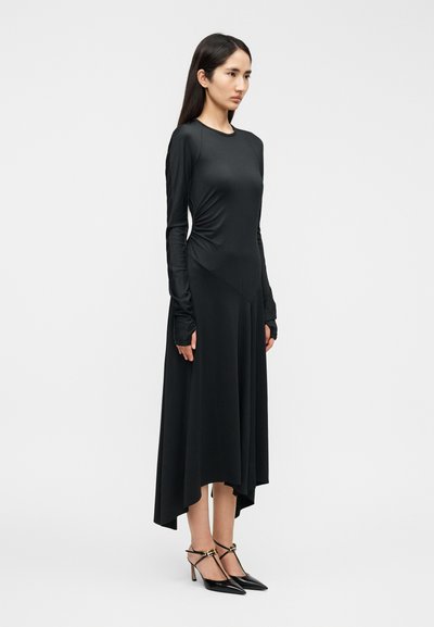 Μαύρο φόρεμα με μακριά μανίκια,  εφαρμοστό μπούστο και ρέον ασύμμετρο τελείωμα. Ομαλή υφή, συνοδευόμενο από μαύρα ψηλοτάκουνα παπούτσια με μυτερή μύτη και χρυσές λεπτομέρειες.