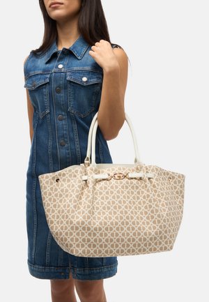 Bolso tote de color beige con un patrón, asas blancas y detalles dorados, sostenido por una persona que lleva un vestido de mezclilla con detalles de botones.