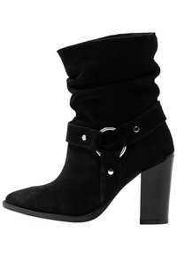 Musta mokkanahka nilkkuri, jossa on löysä yläosa, teräväkärkinen muotoilu ja leveä hihna, jossa on hopea ympyräsolki ja koristepultteja. Paksu korko.