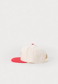 Căciulă de baseball cu coroana crema și viziera roșie. Prezintă o eticheta cu logo pe față, o curea ajustabilă la spate și țesătură texturată.