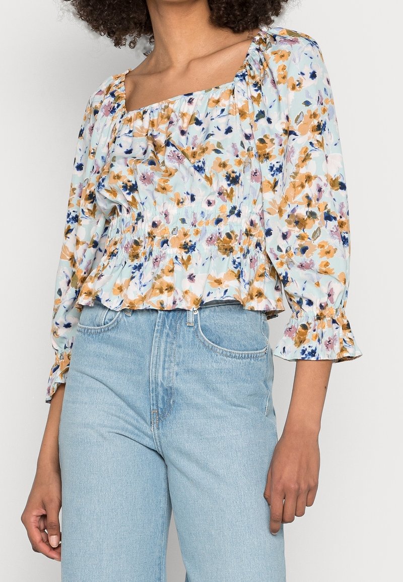 Blouse fleurie avec un fond bleu clair, ornée de fleurs orange et violettes, manches bouffantes et taille froncée. Associée à un jean en denim clair.