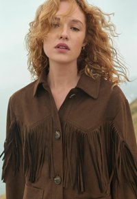 Brown suede jacket featuring fringe detailing along the front and shoulders, large collar, and button closure, set against a soft background.