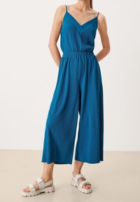 Combinaison côtelée bleu-vert avec un col en V, des fines bretelles, une taille élastique et un design à jambes larges. Associée à des sandales à plateforme grises claires.