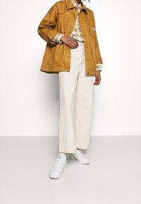 Brun lätt jacka med röda accenter, rutigt skjorta under, krämfärgade vida byxor och vita sneakers. Enkel och funktionell design.