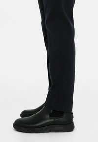 Melni slip-on apavi ar gludu ādas apdari, elastīgām sānu malām un strukturētu gumijas zoli, uzvilkti ar tumšiem biksēm.
