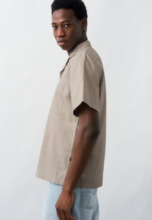 Νεαρός άντρας με μπεζ κοντομάνικο πουκάμισο και ανοιχτόχρωμα μπλε τζιν, που στέκεται πλάγια και κοιτάζει απευθείας την κάμερα, με φόντο μονόχρωμο φόντο.