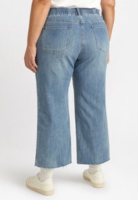 Pantalones anchos de mezclilla azul con cinturilla elástica, que cuentan con bolsillos traseros y dobladillos deshilachados. Combinados con zapatillas blancas y calcetas.