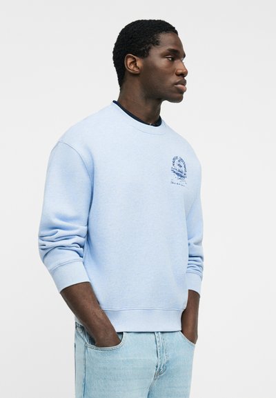 Άνδρας που φοράει ανοιχτό μπλε φούτερ με μικρό σκούρο μπλε γραφικό στο στήθος και ανοιχτό μπλε τζιν, στέκεται με τα χέρια στις τσέπες, κοιτάζοντας δεξιά.