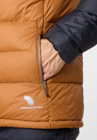 Brun och svart vadderad jacka med en zip-ficka, som har en glänsande yta och "PREMIUM DOWN"-logotyp i vitt. Texturerad, kviltad design.