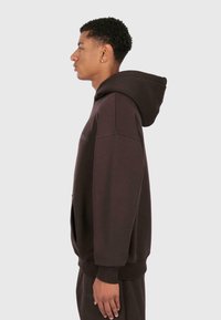 Giovane in una felpa cappuccio marrone scuro e pantaloni abbinati, in posizione di profilo su uno sfondo chiaro e semplice.
