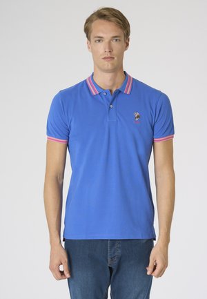 Uomo che indossa una polo blu con colletto e maniche a righe rosa, logo ricamato sul petto, abbinata a jeans blu, in piedi davanti a uno sfondo bianco.