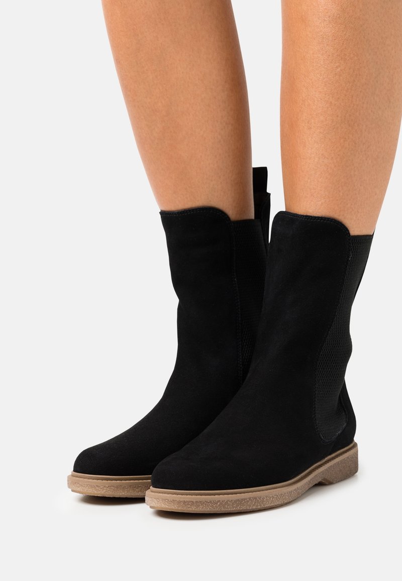 Bottes mi-mollet en daim noir avec semelles en caoutchouc couleur camel, portées sur jambes nues sur fond gris clair.