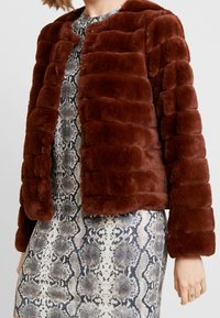 Veste en fausse fourrure marron avec un design matelassé, un col arrondi et des manches longues, portée sur une robe à motifs serpentins.