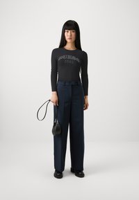Top nero a maniche lunghe con la scritta "GUESS 1981" in lettere grigie, abbinato a pantaloni cropped blu scuro a vita alta e una piccola borsa nera.