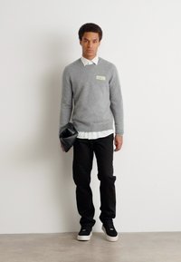 Pilkas megztinis ant balto marškinių sukriaustų, juodos kelnės ir juodi batai. Laiko juodą rankinę. Vienodiniai spalvos, minimalistinis dizainas.