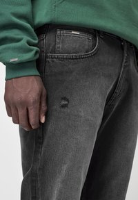 Närbild av en mörkhyad hand som vilar bredvid bleka svarta jeans med ett litet hål nära fickan, ihop med en grön långärmad sweatshirt.