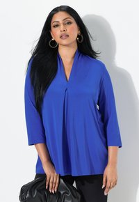 Top blu, a maniche lunghe, con scollatura a V e dettagli a plissettatura, realizzato in tessuto morbido. Abbinato a una borsa nera.