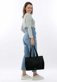 Zwarte gestructureerde tote bag met twee handvatten, vastgehouden door een persoon in een gestreepte lange mouwen top en high-waisted denim jeans.