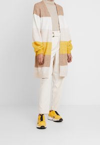 Sweter-kardigan z pionowymi paskami w beżu, bieli i żółci, z długimi rękawami i otwartym krojem, zestawiony z kremowymi spodniami i żółtymi tenisówkami.