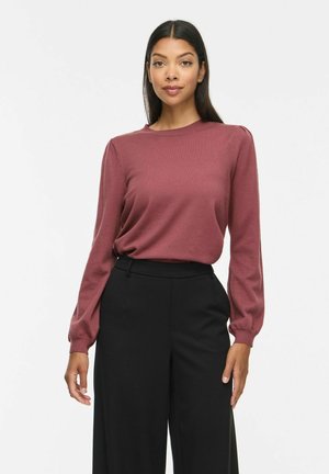 Femme aux longs cheveux foncés portant un haut mauve à manches longues et un pantalon noir taille haute à jambes larges, debout sur un fond blanc.