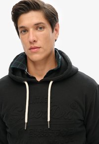 Sudadera negra con logo texturizado, cordones en color crema, superpuesta sobre una camisa de cuadros verde y azul. Fondo simple con tonos neutros.