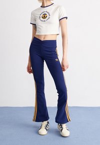 Zkrácený bílý tričko s modrým lemem a grafikou, doplněné námořnickými zvonovými kalhotami s žlutými pruhy a vyšitým logem. Texturovaná látka.