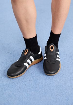 Jambes portant des chaussettes noires et des chaussures en cuir noir avec des rayures blanches et une semelle en gomme, sur un sol texturé bleu clair.