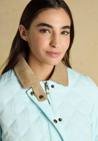 Mujer joven con cabello largo y oscuro que lleva una chaqueta acolchada azul claro con cuello de pana beige, mirando con una ligera sonrisa frente a un fondo beige.