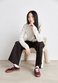 Femme aux longs cheveux noirs portant un pull blanc, un pantalon large noir et des baskets bordeaux, assise sur une chaise beige moderne contre un mur blanc.