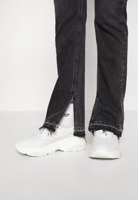 Zapatillas blancas gruesas combinadas con jeans de mezclilla oscura que tienen dobladillos deshilachados y una abertura en el tobillo, que revela calcetines blancos debajo.