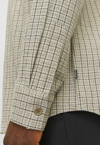 Chemise beige à boutons avec un motif à carreaux rectangulaires en noir. Présente des manches à poignet avec un bouton circulaire. Tissu texturé.