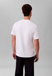 Biały bawełniany t-shirt z okrągłym dekoltem i krótkimi rękawami, widziany od tyłu, zestawiony z ciemnymi dżinsami. Prosty fason, bez wzorów i ozdobników.