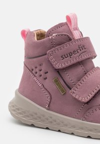 Lilla semsket høyhælte sko med to borrelåsremmer, Gore-Tex-merke, teksturert gummisåle og rosa detaljer på hælen og remmen.