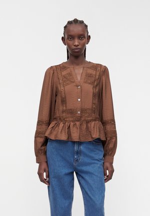 Frau mit geflochtenem Haar, die eine braune langärmelige Spitzenbluse mit Knöpfen, Peplum-Saum und blaue hoch taillierte Jeans trägt, steht vor weißem Hintergrund.