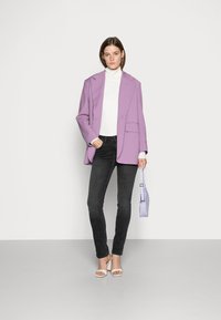 Blazer oversized lilás, suéter de gola alta branco em padrão canelado, jeans skinny pretos, sandálias de salto brancas e uma pequena mala lavanda. Textura suave, ajuste feito à medida.