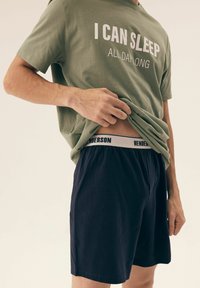 Zielona koszulka bawełniana z białym napisem "MOGĘ SPAĆ PRZEZ CAŁY DZIEŃ", w połączeniu z granatowymi szortami z białym pasem oznaczonym "HENDERSON".