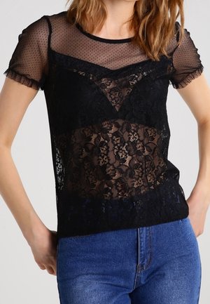 Frau trägt ein schwarzes, kurzärmeliges, transparentes Spitzentop mit Blumenmustern über einem Top, kombiniert mit blauen Jeans.