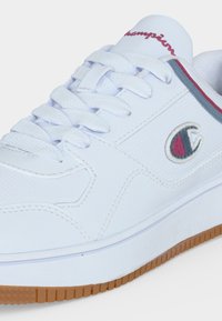Baskets basses blanches avec lacets blancs, semelle en gomme, logo Champion rouge et bleu marine sur le côté, et texte Champion rouge sur la languette.