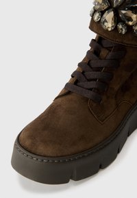 Stivale in suede marrone con suola nera spessa, caratterizzato da lacci scuri e impreziosito con gemme di colore scuro nella parte superiore.