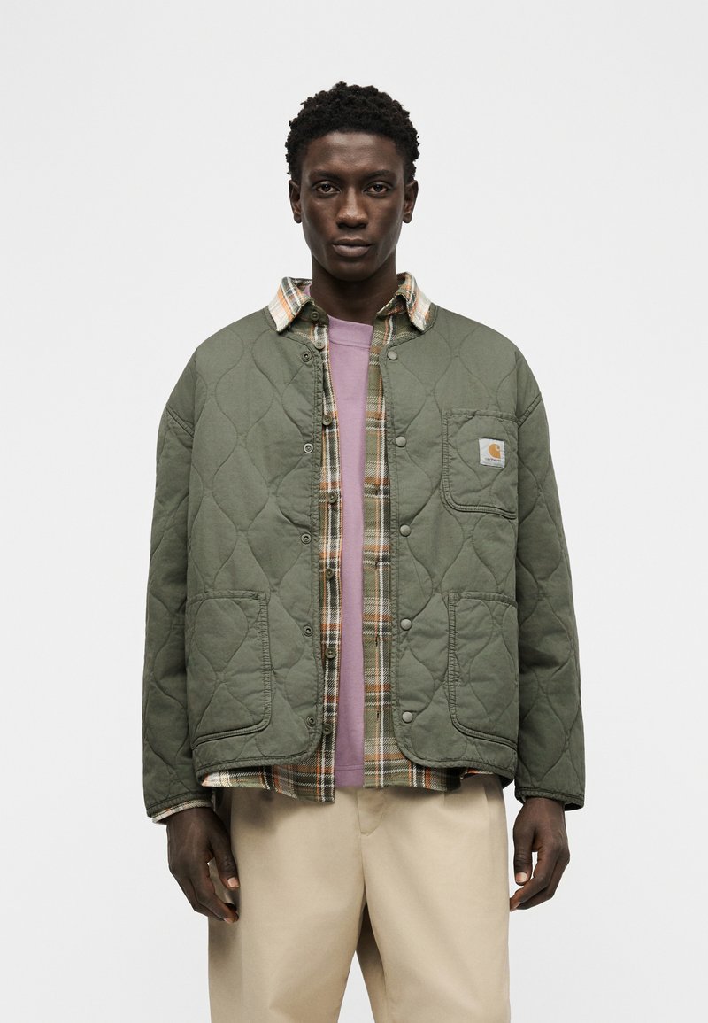 Jeune homme portant une veste matelassée verte par-dessus une chemise à carreaux et un t-shirt rose, associé à un pantalon beige sur un fond blanc uni.