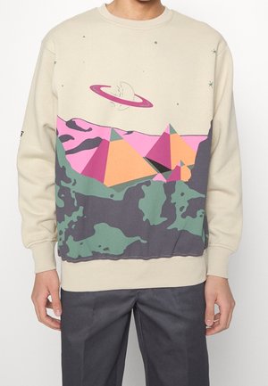 Beigefarbener Pullover mit abstrakten geometrischen Bergen in Pink, Orange und Grün, mit einem gesprungenen Planeten mit einem Ring und kleinen Sternen darüber.