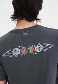 Camiseta gris con gráficos coloridos de rosas y alas en la parte posterior, con una tela texturizada y una pequeña etiqueta metálica de la marca en el cuello.
