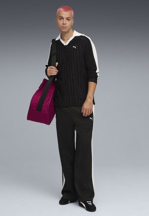 Czarny pulower z fakturą i białymi akcentami, czarne spodnie dresowe z białymi paskami oraz magentowa torba podróżna, trzymana przez osobę z różowymi włosami.