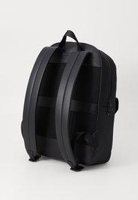 Sac à dos noir doté de doubles sangles rembourrées, d'un dos en maille texturée et d'une poignée supérieure. Fermeture éclair avec une surface lisse et un design minimaliste.