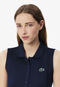 Polo sans manches bleu marine avec un col, présentant une patte de boutonnage et un logo crocodile vert sur le côté gauche. Texture lisse.