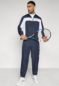 Tumši zils sporta tērps ar baltām detaļām, rāvējslēdzēja jaka un elastīgi aproces, kopā ar tenisa raketi un baltām sporta kurpēm.