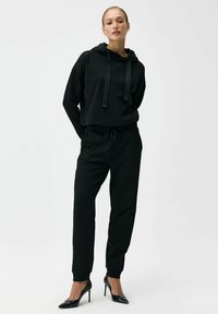 Czarna bluza z kapturem z troczkami, zestawiona z czarnymi spodniami typu jogger, oba wykonane z miękkiego materiału. Noszone z czarnymi szpilkami. Prosty design.