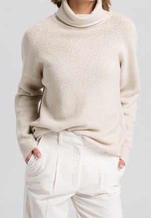 Woman wearing a cream knit turtleneck sweater with textured pattern and loose off-white high-waisted pants, hands in pockets.