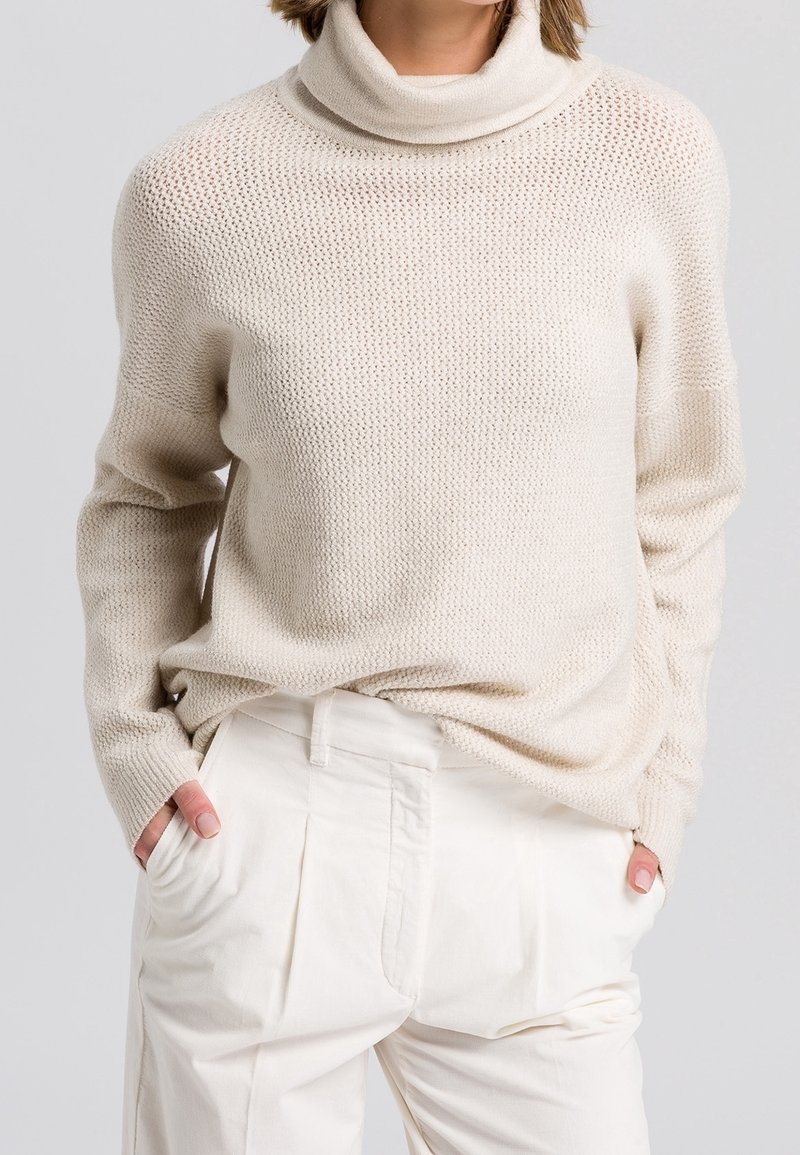 Femme portant un pull à col roulé en tricot crème avec un motif texturé et un pantalon taille haute ample blanc cassé, les mains dans les poches.